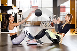 Two young athletic girls dressed in sportswear are doing together sport exercises for press with fitness ball in the