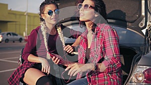 Two young attractive girlfriends in sunglasses sitting inside of the open car trunk in the parking by the shopping mall