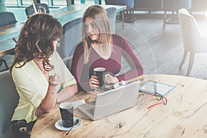 Two young attractive women sit at round wooden table in cafe, drinking coffee and talking. Business meeting.