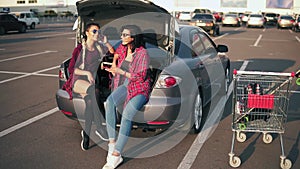 Two young attractive women in sunglasses sitting inside of the open car trunk and listening to the music in the