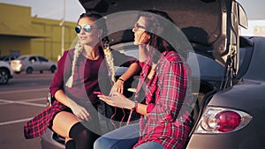 Two young attractive women in sunglasses sitting inside of the open car trunk in the parking by the shopping mall and