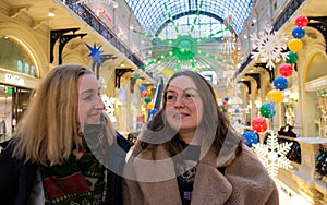 Two young beautiful female friends at the shopping mall smiling, admire the bright Christmas decor