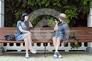 Two young beautiful girls in jeans dresses and hats sit on a bench in the park on a background of green plant walls, and