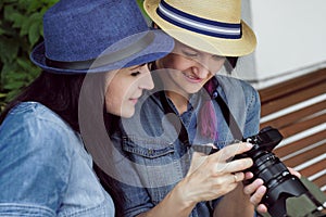 Two young beautiful girls in jeans dresses and hats sit on a bench in the park on a background of green plant walls, and