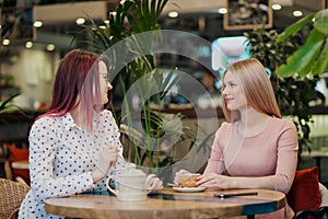 Two young beautiful girls sit in a green cafe at a table. Drink tea with croissants, chatting, laughing and taking