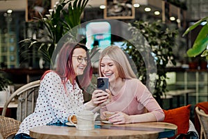 Two young beautiful girls sit in a green cafe at a table. Drink tea with croissants, chatting, laughing and taking