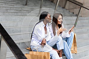 Two young beautiful girls use phone while sitting on the mall stair with shopping bags