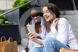 Two young beautiful girls use phone while sitting on the mall stair with shopping bags