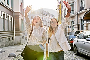 Two young beautiful happy women university students walking along city street