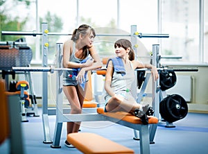 Two young beautiful slim women in grey sportswear with towels with bottle of water and looking at each other in gym