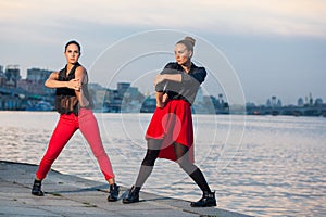 Two young beautiful twin sisters are dancing waacking dance in the city background near river.