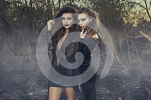 Two young beautiful witches in black gowns standing in the middle of burnt meadow with predatory face expression.