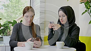 Two young beautiful womans sitting in cafe and typing on phones, one of them muslim woman in hijab, determined and