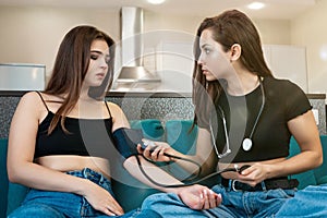 Two young beautiful women friends sitting on the sofa, one girl is sick, her friend helps her to take blood pressure using