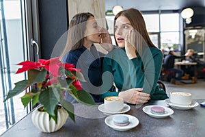 Two young beautiful women secretly sitting at a table in a coffee shop, girls whisper a secret in their ear