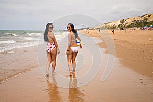 Two young and beautiful women stroll along the beach. One of them is carrying a volleyball. The girls are wearing bikinis and