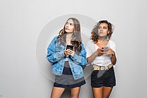 Two young best friends women are busy at their mobile phones in social nets, wearing casual clothes, isolated on white background