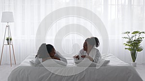 Two Young Black Sisters Wearing Bathrobes Relaxing On Bed At Home
