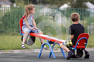 Two young blond children preschooler girl with long ponytail and cute school boy swing on see- saw on bright green blurred backgro