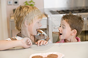 Two young boys in kitchen eating cookies smiling