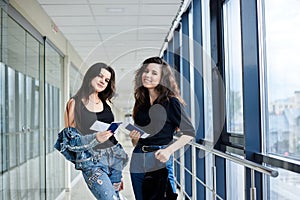 Two young brunette girls, standing in light airport hallway with huge windows, wearing casual jeans clothes, holding international