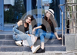 Two young brunette girls, wearing casual jeans attire, sitting on stairs of blue modern glass building. Girlfriends traveling in