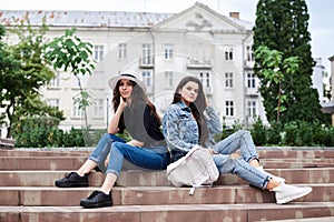 Two young brunette girls, wearing casual jeans clothes, holding small backpacks, sitting on stairway, resting, in front of