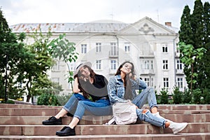 Two young brunette girls, wearing casual jeans clothes, holding small backpacks, sitting on stairway, resting, in front of
