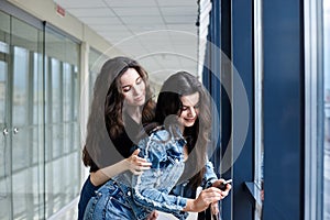 Two young brunette girls, wearing casual jeans clothes, leaning on railing in light airport hallway with huge windows, checking