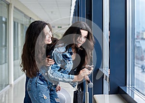 Two young brunette girls, wearing casual jeans clothes, leaning on railing in light airport hallway with huge windows, checking