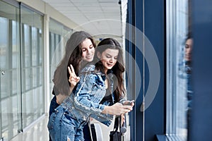 Two young brunette girls, wearing casual jeans clothes, leaning on railing in light airport hallway with huge windows, checking