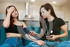 Two young brunette women friends sitting on the sofa, one girl feels sick, her friend helps her to take blood pressure using