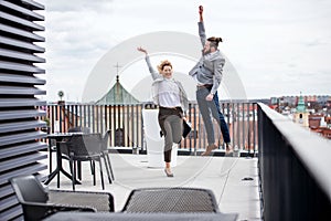 Two young business people jumping on terrace outside office, expressing excitement.