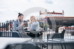 Two young business people with laptop sitting on a terrace outside office, laughing.