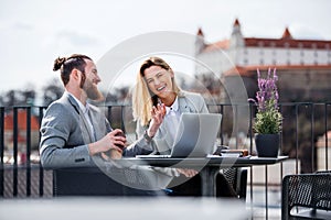 Two young business people with laptop sitting on a terrace outside office, working.