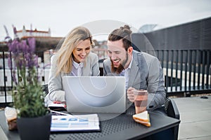Dvaja mladí podnikatelia s notebookom sedia na terase mimo kancelárie, pracujú.