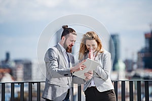 Two young business people with tablet standing on a terrace outside office, working.