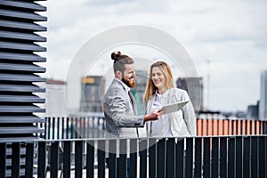 Two young business people with tablet standing on a terrace outside office, working.