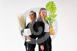 Two young business woman dressed black suit smile holding houseplant and lamp standing isolated on white background. Moving