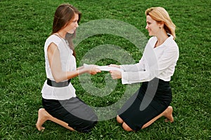 Two young business women are fighting for a stack of blank papers against the backdrop of green grass. Outdoor photo