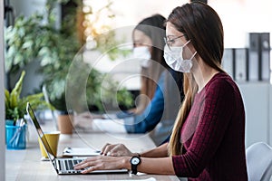 Two young business women friends wearing a hygienic face mask while work with laptops in the coworking space