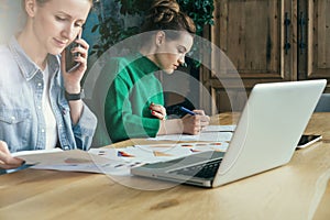 Two young business women sitting in office at table and work together.On table laptop and paper charts.
