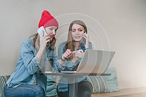 Two young business women sitting at table in cafe and talking on cell phone,while watching on screen laptop