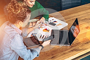 Two young business women sitting at table in front of laptop.On table is tablet computer and paper charts.