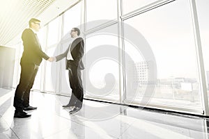 Two young businessmen are shaking hands with each other standing against panoramic windows background