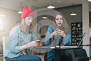 Two young businesswomen sitting at table in coffe shop, look at your smartphone screen and discuss business strategy