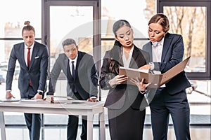 Two young businesswomen working together with folder while businessmen standing behind
