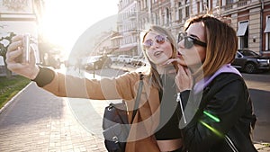 Two young caucasian fashion women in sunglasses posing for selfie and laughing with tonque. Lifestyle portrait on street