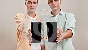 Two young caucasian twin brothers in casual wear holding smartphone with blank screen while standing isolated over beige