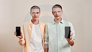 Two young caucasian twin brothers in casual wear holding smartphone with blank screen while standing isolated over beige